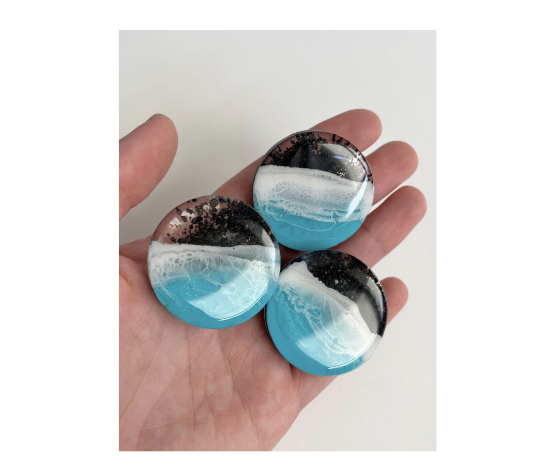 a hand holding out 3 round resin magnets, crafted in a dark brown, white a turquoise pattern that looks like waves washing up on a beach.