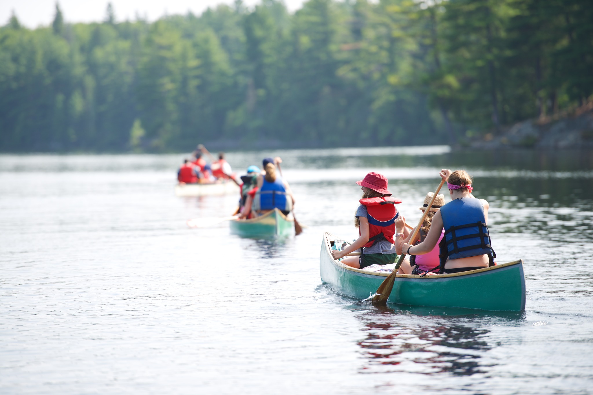 a line of canoes filled with people of all ages paddle into the distance along a silvery river skirted by dense green forest on a summer day.