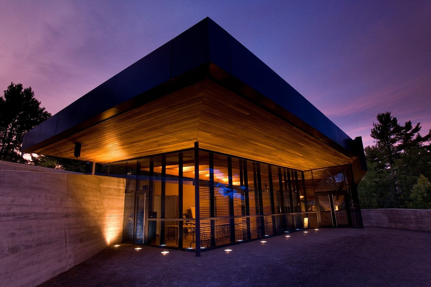 The French River Visitor Centre; A stately wooden structure with high peaked roof and glass foyer, illuminated in the evening. Behind it is the dark silhouettes of trees and a purple and pink twilight sky.