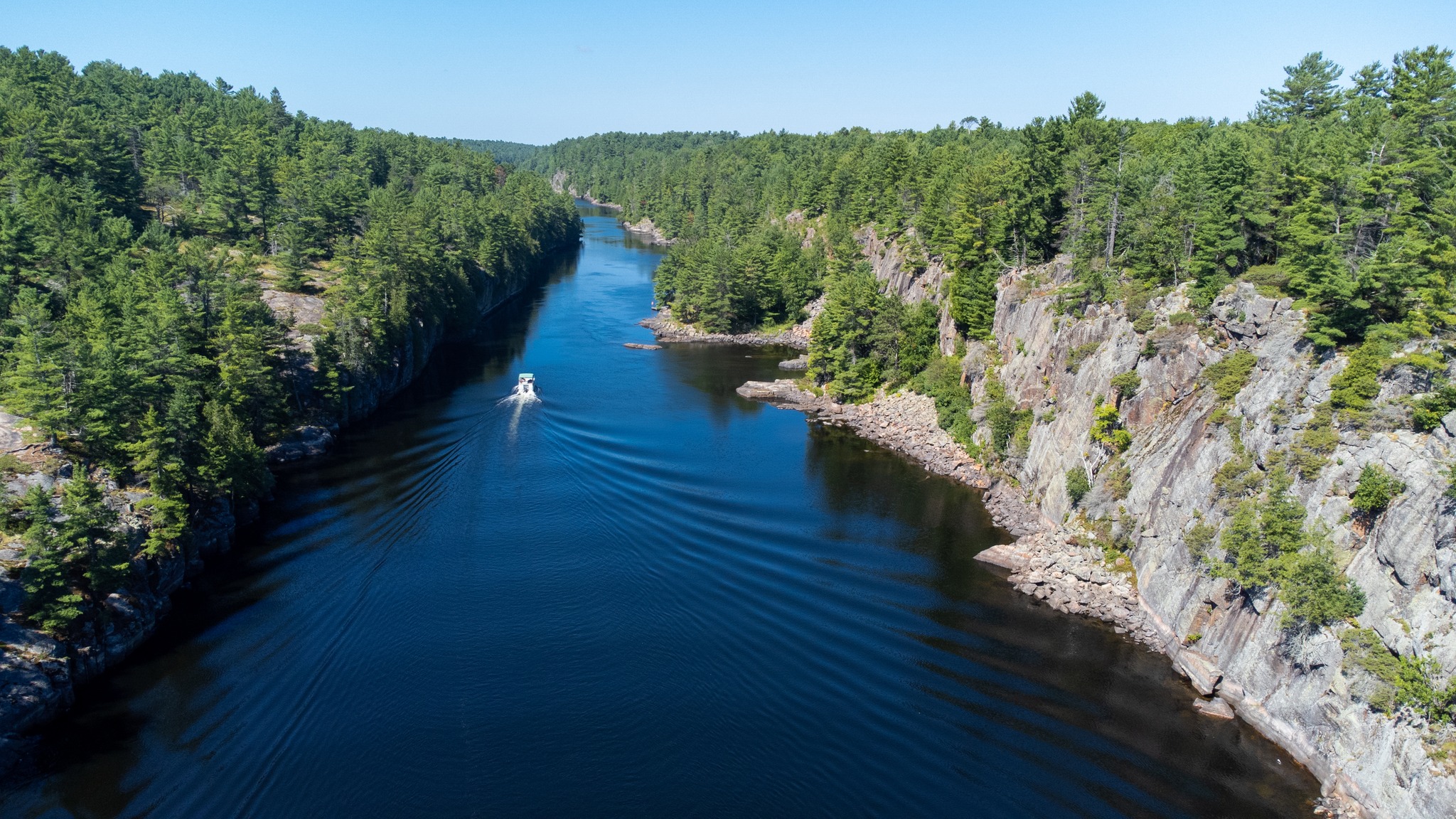 An aerial shot of of a small boat sailing down the wide, calm blue French River, bordered by steep rock banks and boreal forest, under a clear blue sky.