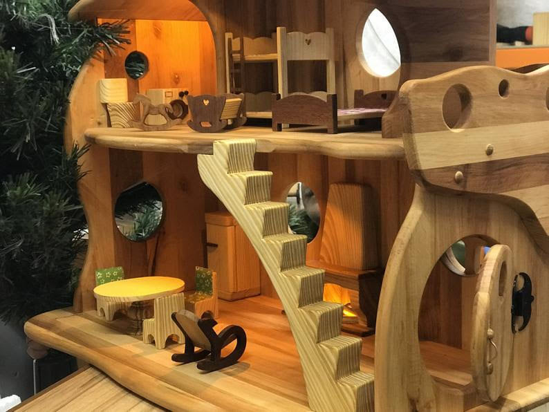 a bi-level wooden dollhouse with a tiny staircase that leads to a nursery with wooden bunkbeds and a cradle. On the lower floor are rocking chairs, a table, circular windows and doors that open. It sits next to a Christmas tree.