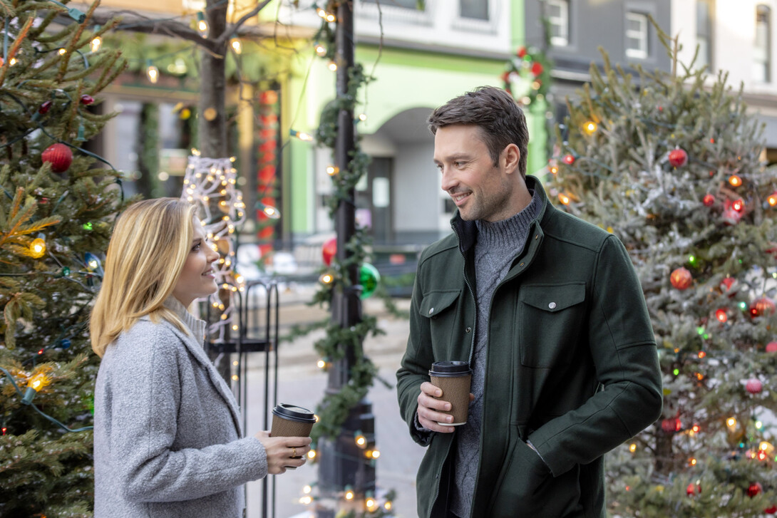 A white couple acting in a movie scene with Christmas trees in the background. 