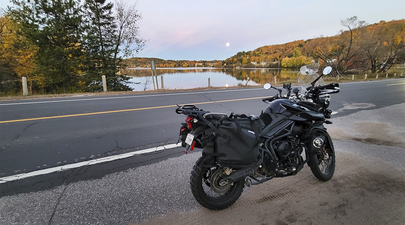 a motorcycle parked at the side of a quiet, paved road next to a calm lake surrounded by autumn forest at sunrise.