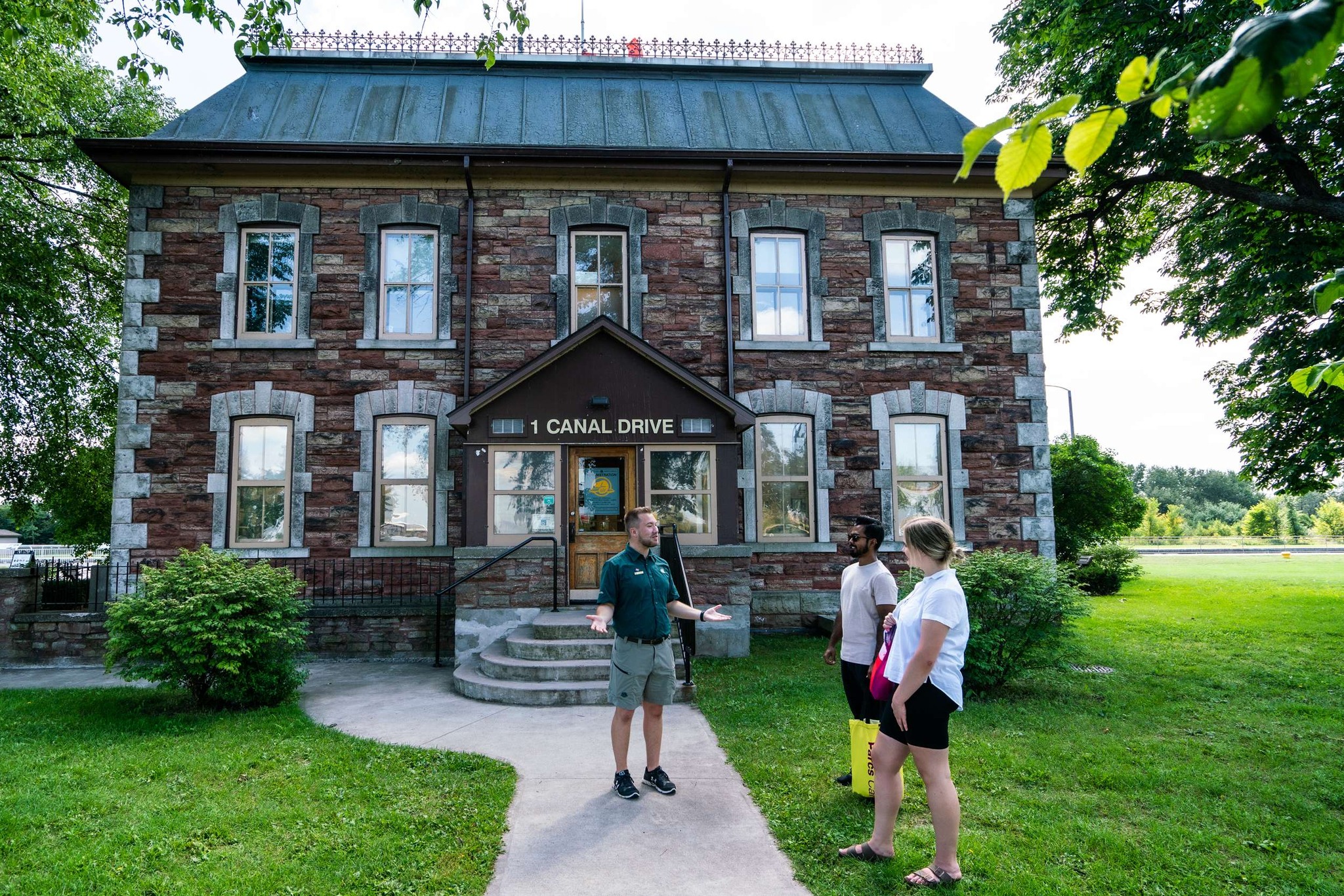 A tour guide talks to two tourists as they stand outside one of the stone historical buildings at the Sault Ste Marie Canal National Historic Site.