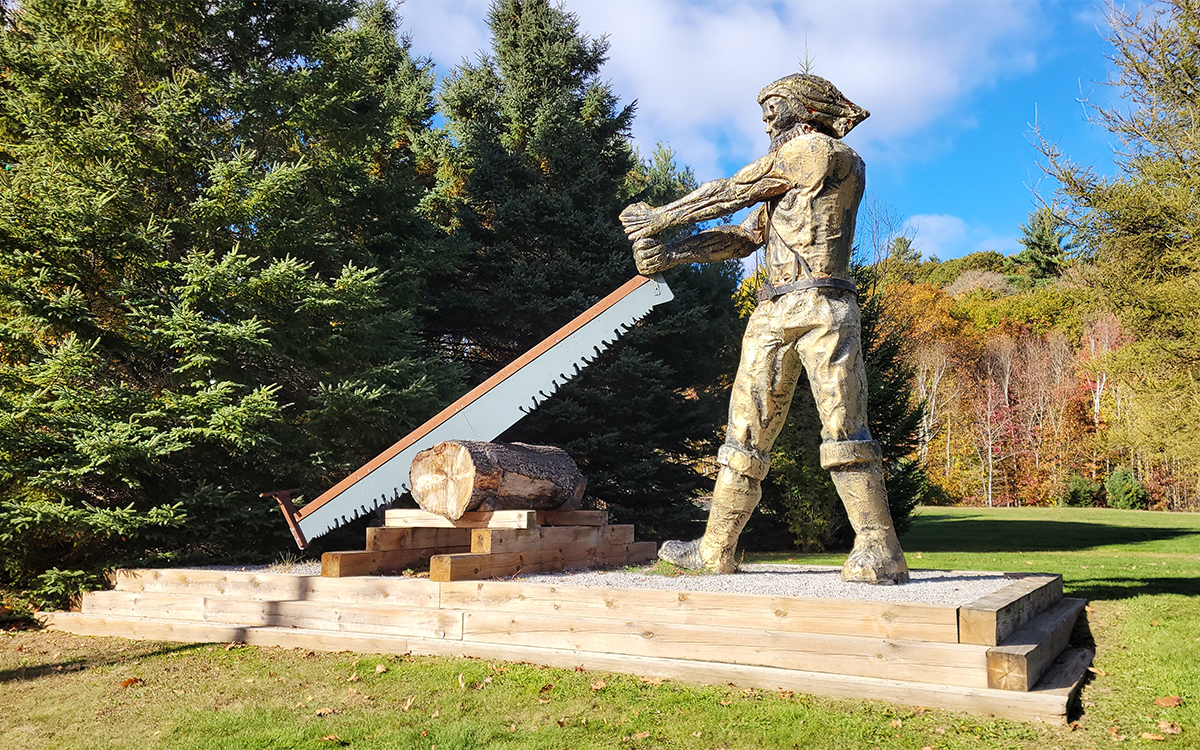 a large carved wooden lumberjack sawing a log, surrounded by trees and grass.