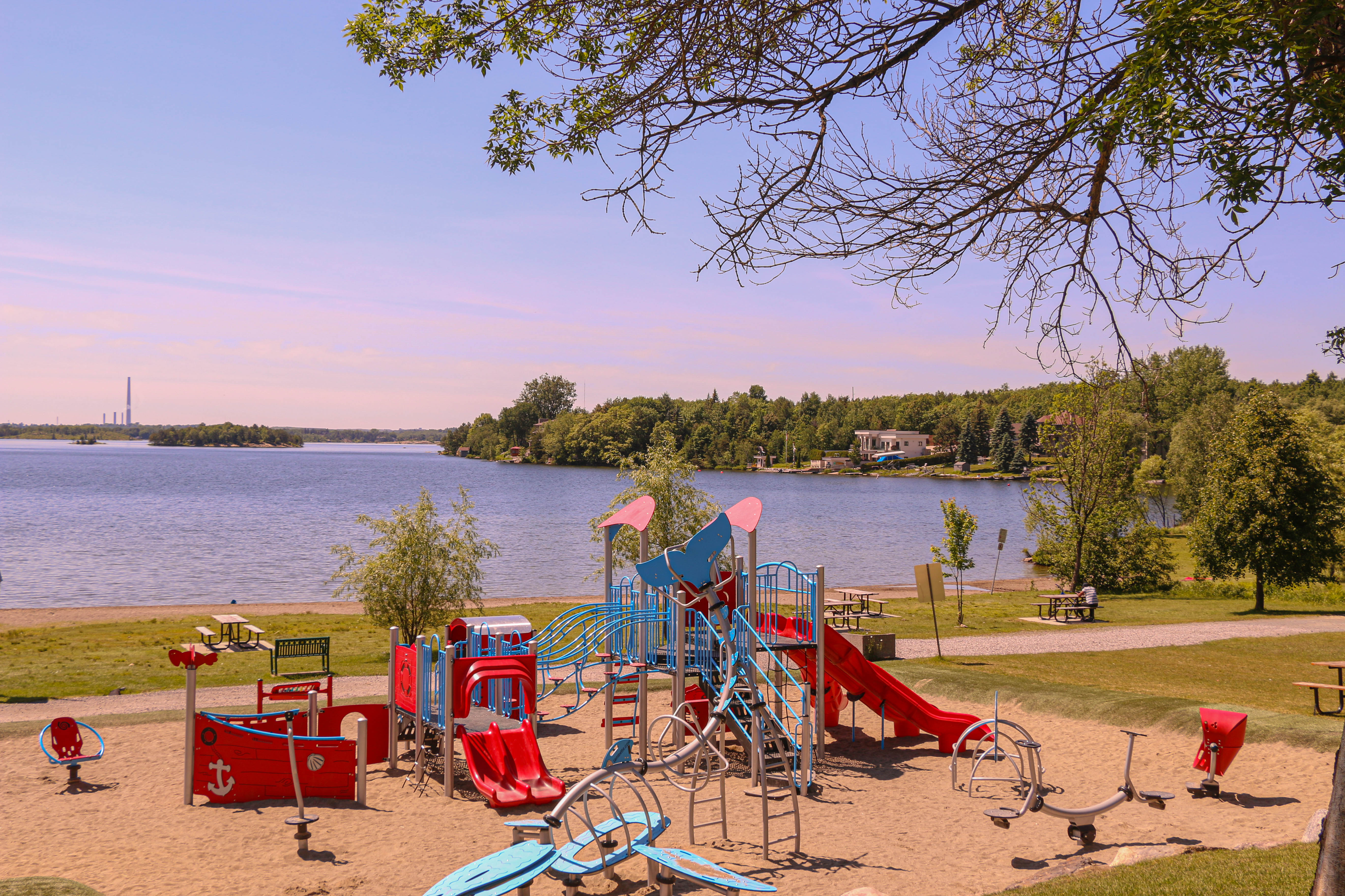 Moonlight Beach; a large playground surrounded by sand, next to a sandy beach by a calm blue lake skirted by green forest. The summer sky is glowing with a light pink and blue sunset. 