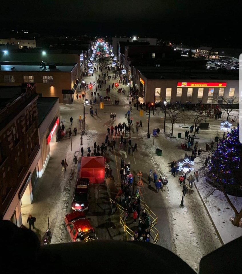 a snowy main street in North Bay far below at night, with a parade of people and sleighs walking down the lit-up street.