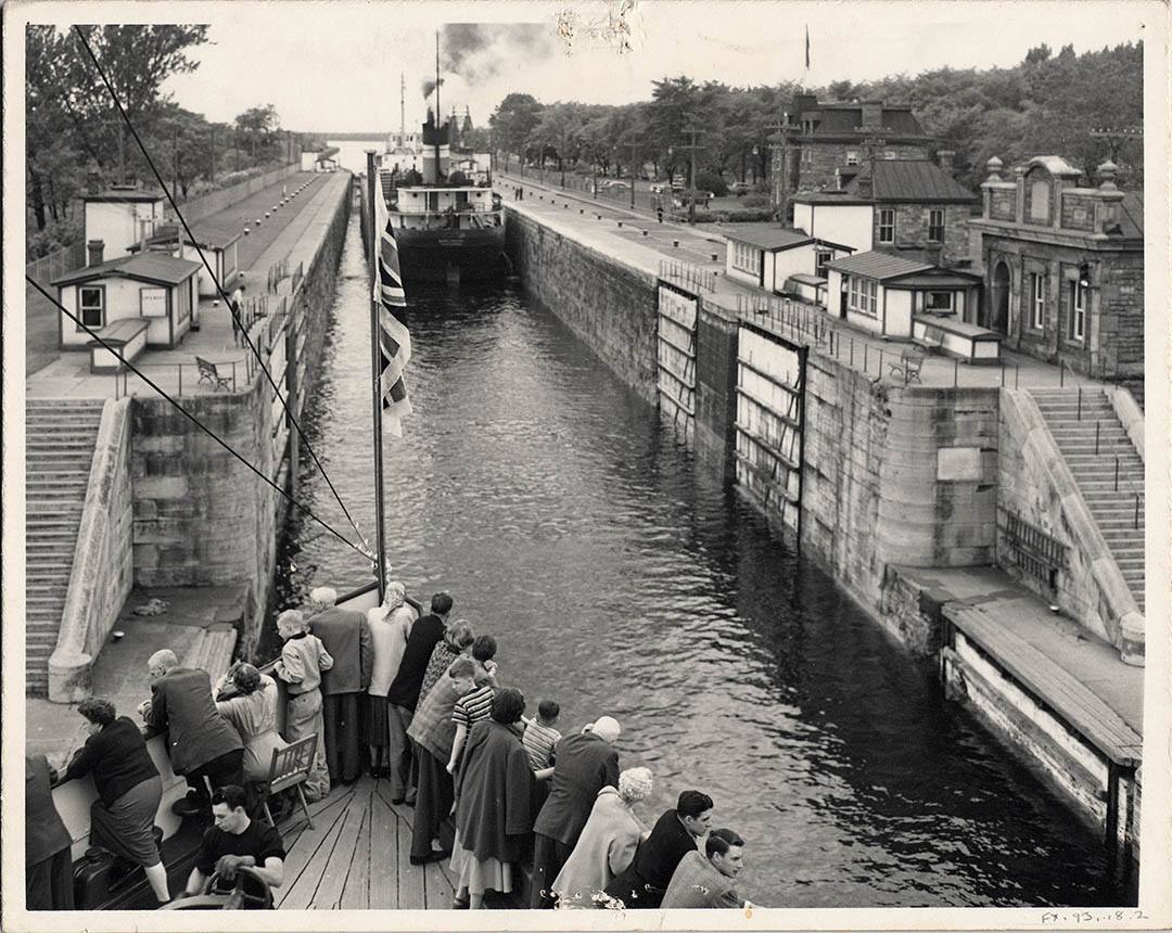 a black and white historical photo of people lined up along the Sault Ste. Marie locks, watching a steamboat come through the canal.