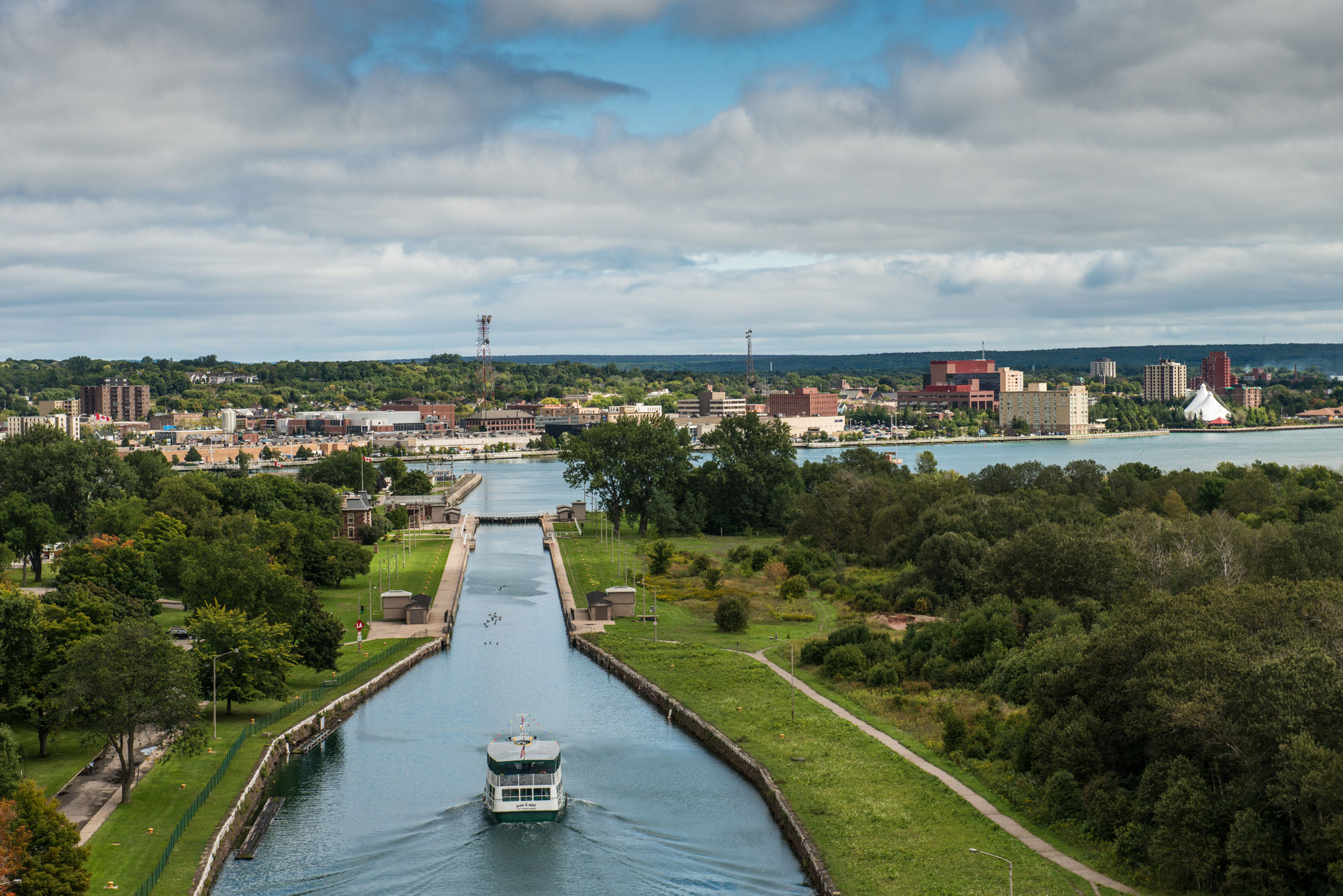 A boat sailing down the Sault Ste. Marie Canal, into the locks. The canal is surrounded by lush green grass and trees and the rest of the city sits on the horizon.