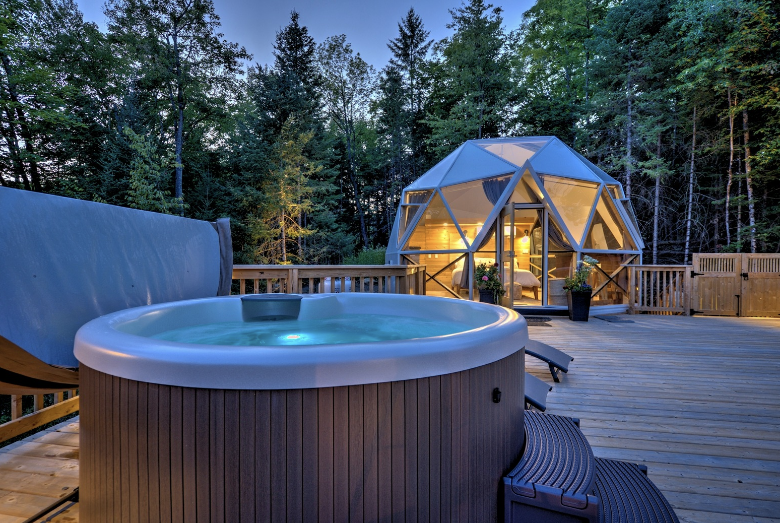 A hot tub and glamping dome on a wooden deck surrounded by pine trees. 