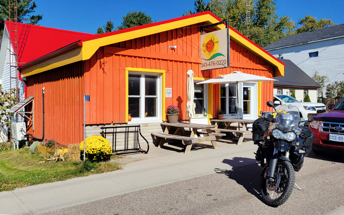 Sun Run Cafe; a small shop with orange wooden siding next to a paved street, with a motorcycle parked in front, on a very sunny day.