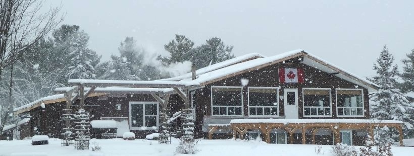 A large wooden cabin with deck next to a lake, covered in a blanket of falling snow. There is forest all around.