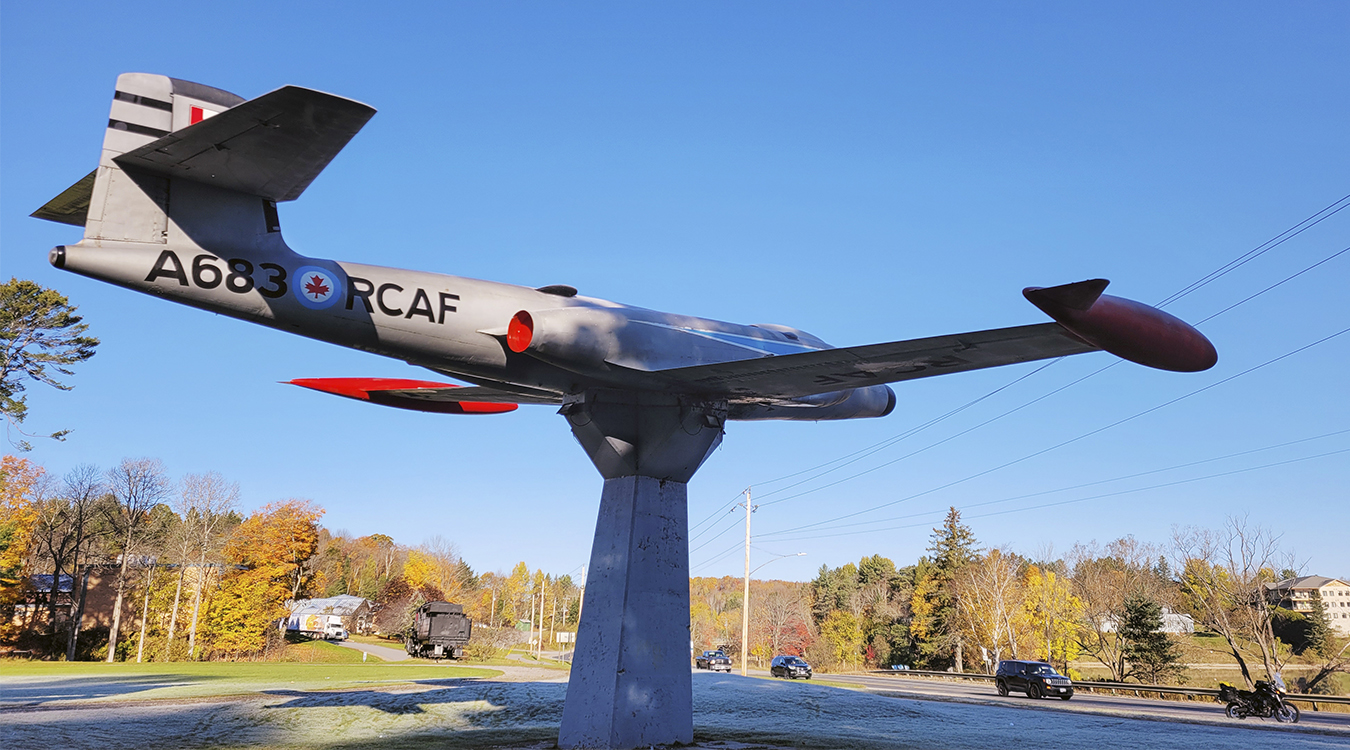 a replica airplane on a stand surrounded by autumn forest, under a blue sky.