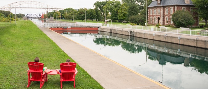 Two people sit holding hands in red Adirondack chairs, looking out over the Sault Ste. Marie Locks at the Sault Ste. Marie National Historic Site.