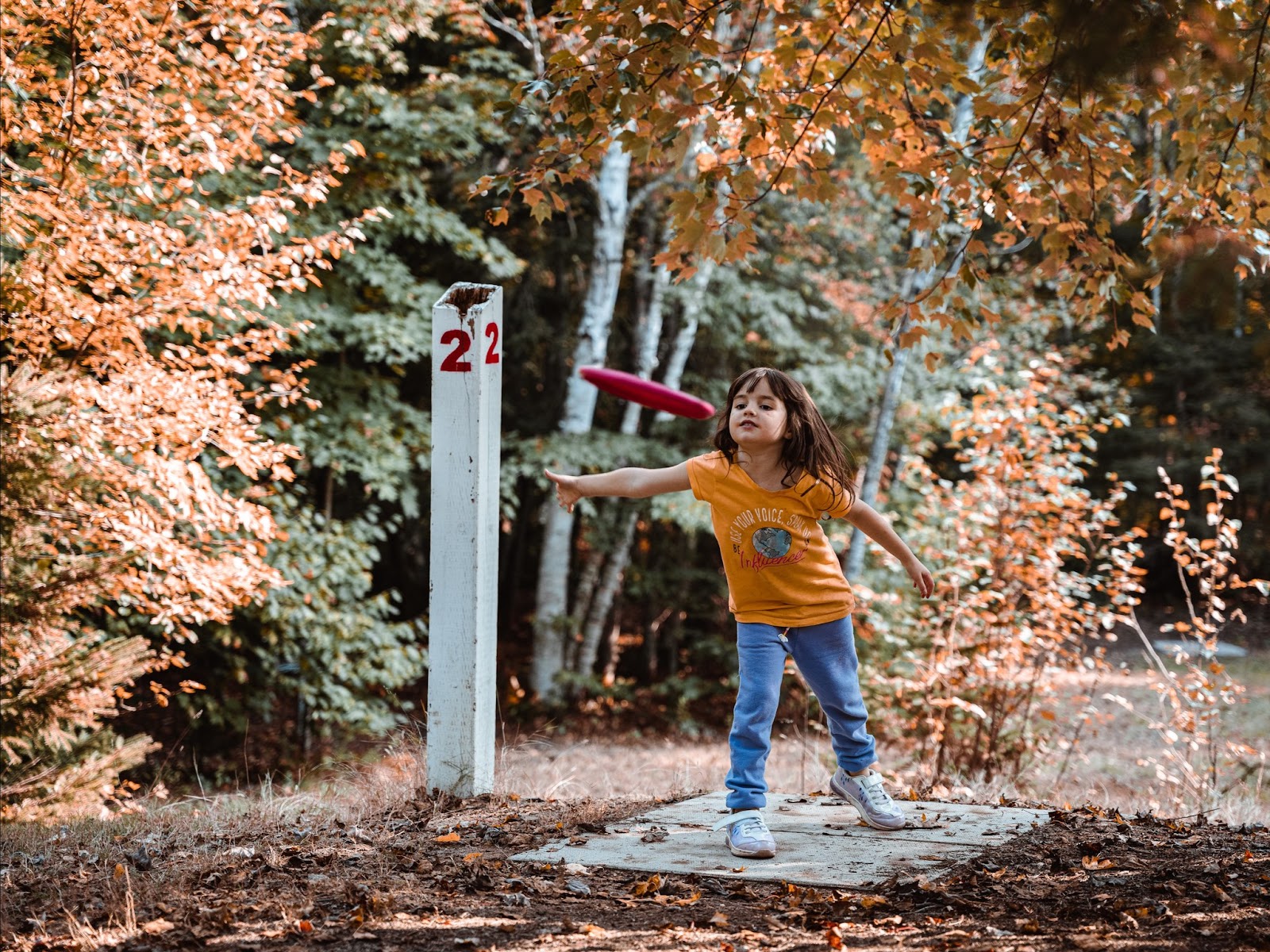 a girl throws a disc golf disc on a course surrounded by forest on a autumn day. 