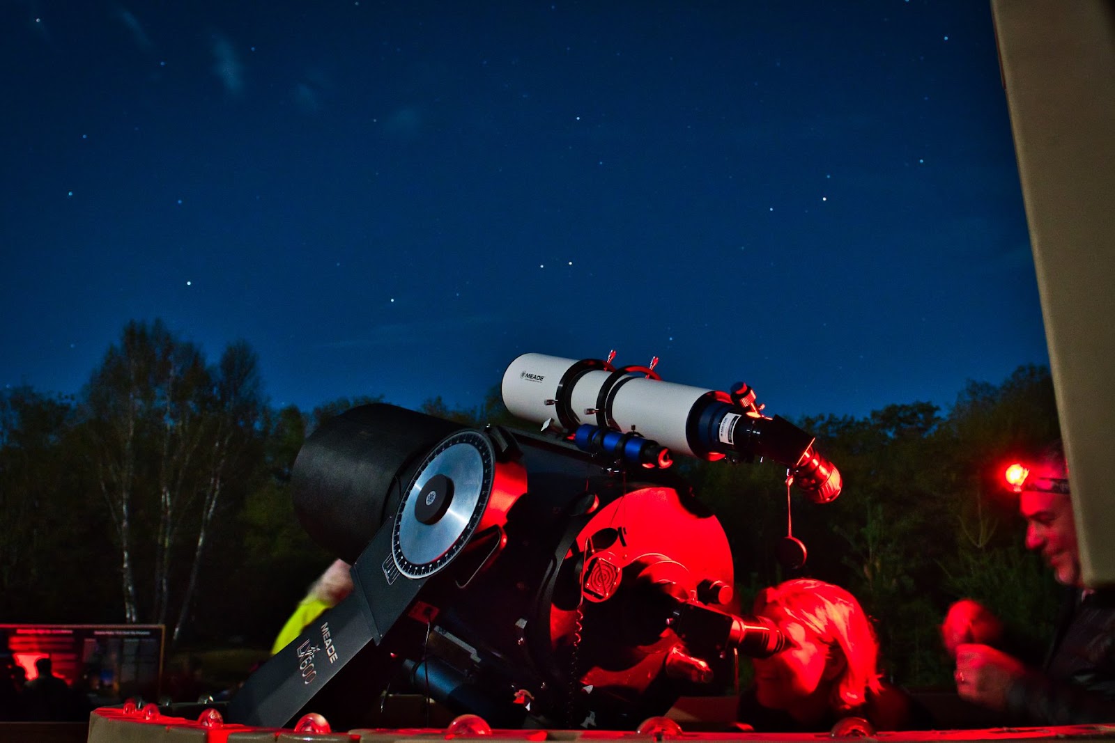 a telescope and bags of gear and stargazing equipment sit in the foreground while two smiling people wearing headlamps ready the telescope under a dark starry sky in a forest.