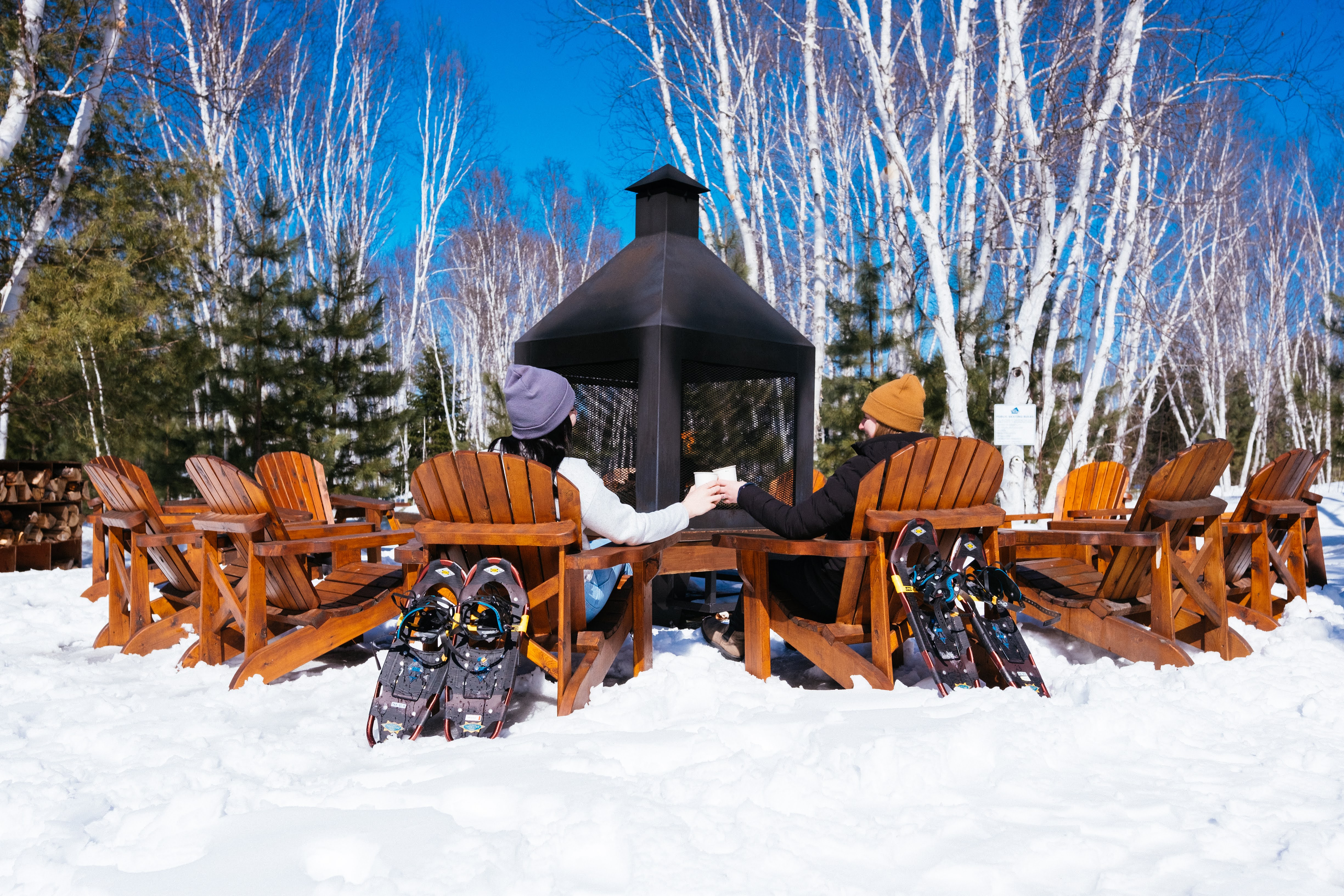 two people in winter clothing clink mugs of hot chocolate together as they lounge in wooden chairs around a fire on a sunny winter day. Shoeshoes are leaning up against their chairs.