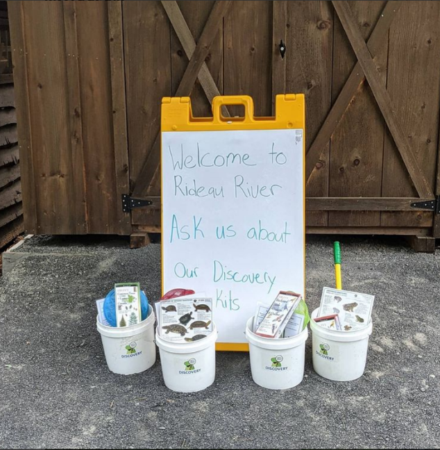 small informational science kits sit next to a stand-up white board which reads "Welcome to Rideau River, ask us about our Discovery Kits".