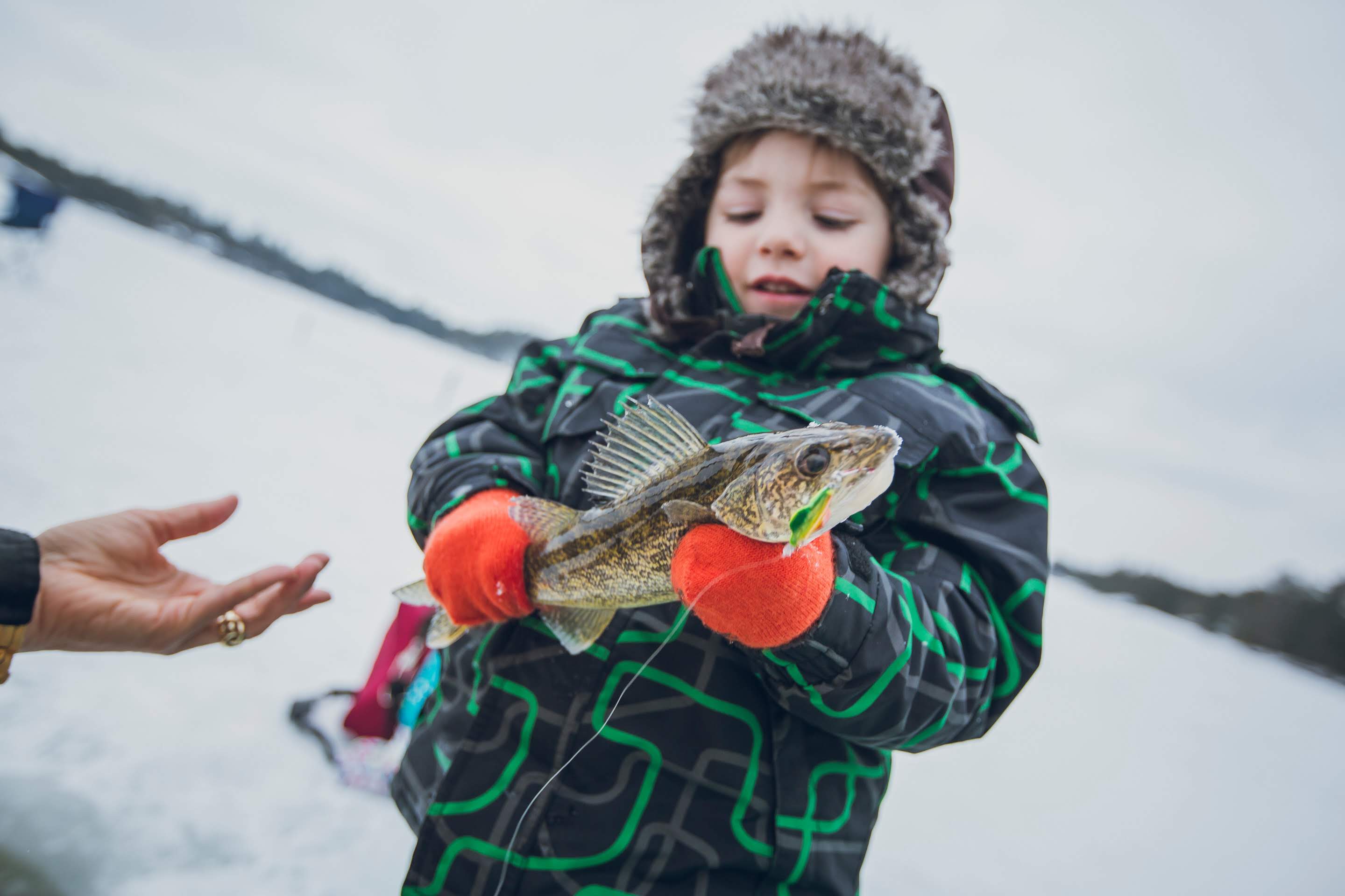 a small boy in winter clothing smiles down at a fish he is holding while standing on a frozen lake.