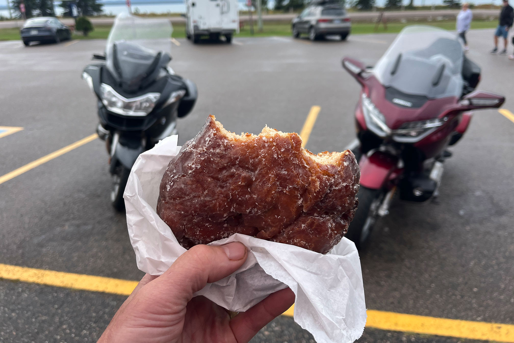 A hand holds up a fritter with bites taken out of it with 2 motorcycles parked in the background.