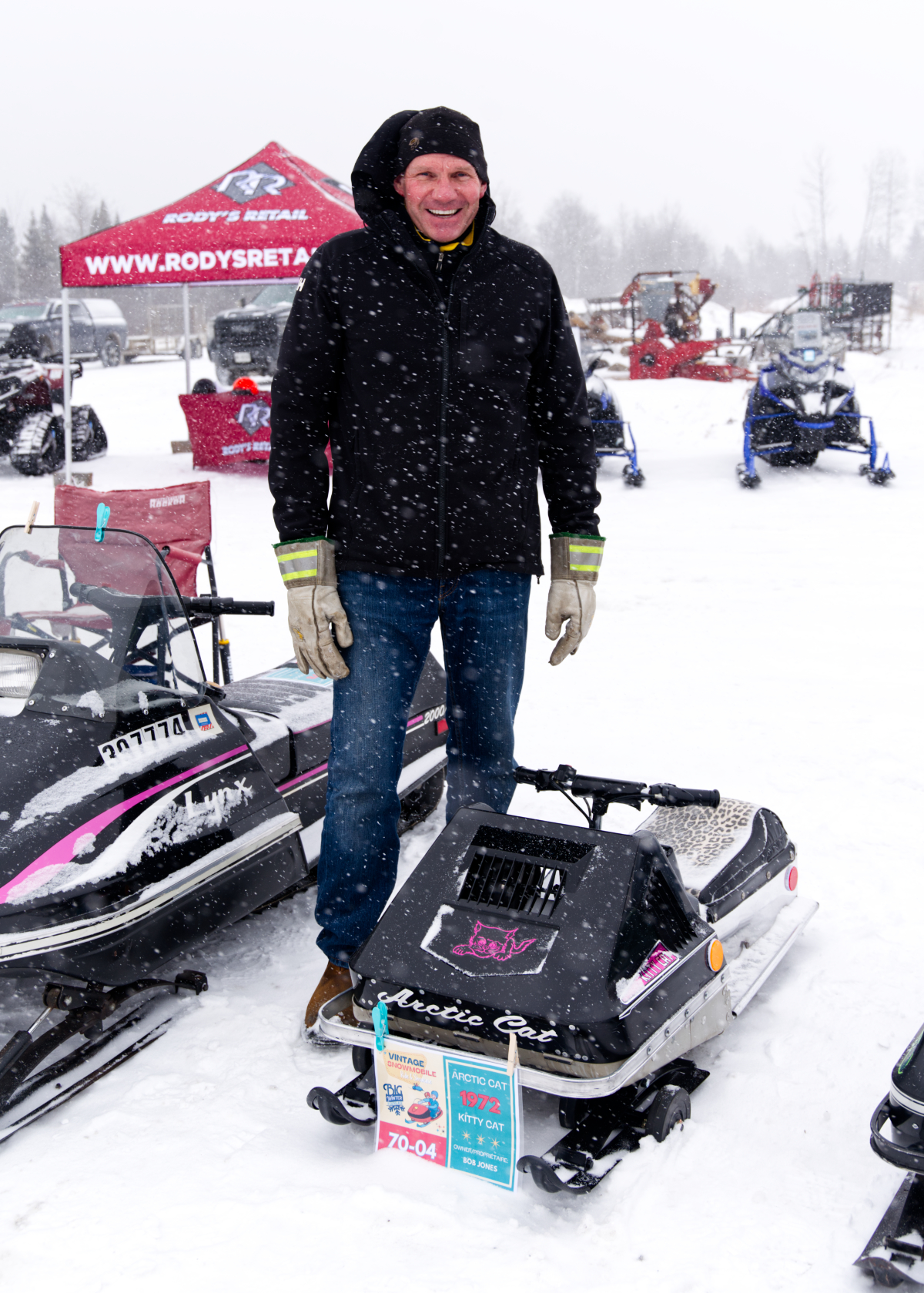 A tall man smiles while standing next to a very tiny vintage snowmobile. 