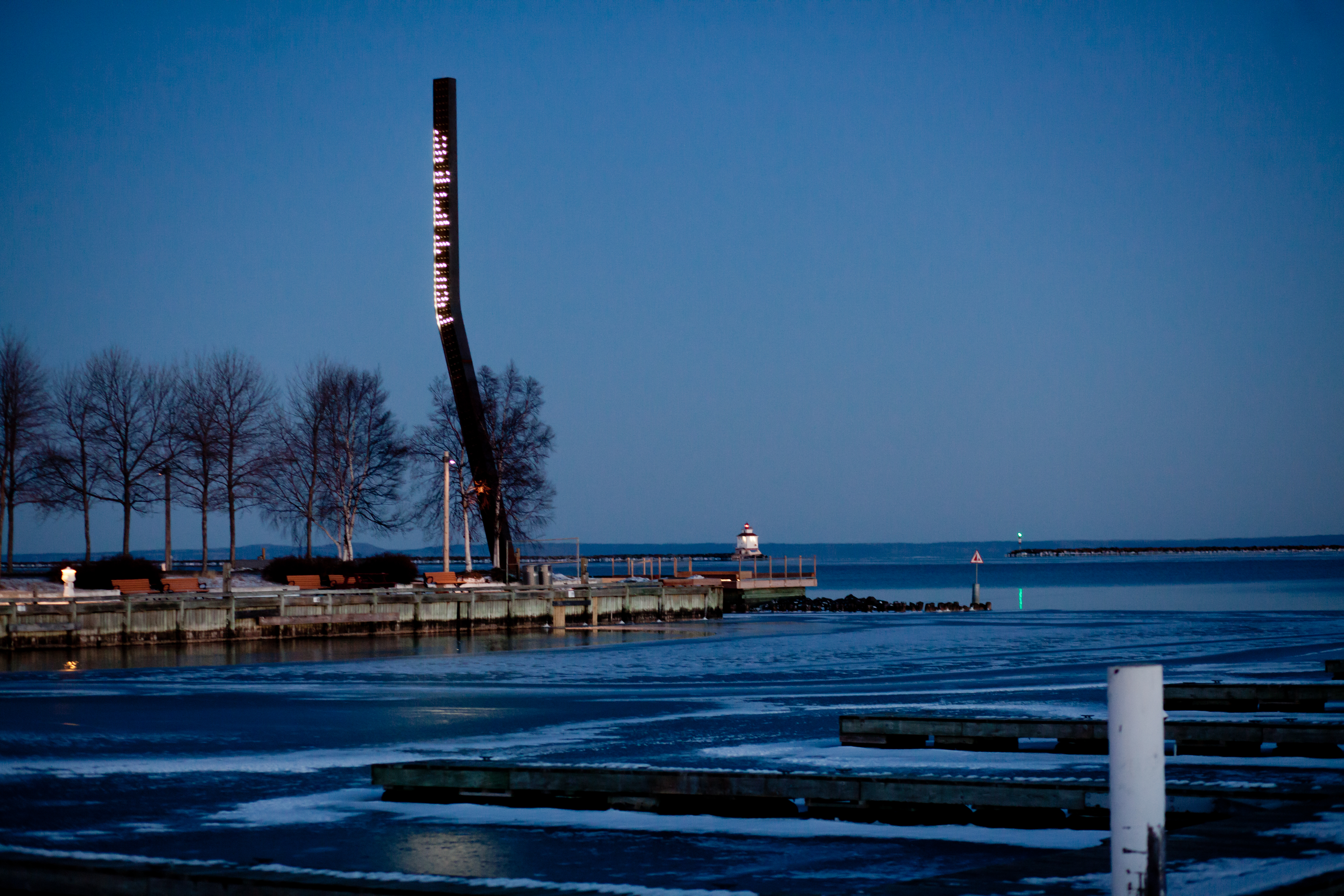 Prince Arthur's Landing; a tall lit beacon towering above the waterfront under a blue twilight. The trees along the waterfront are bare.