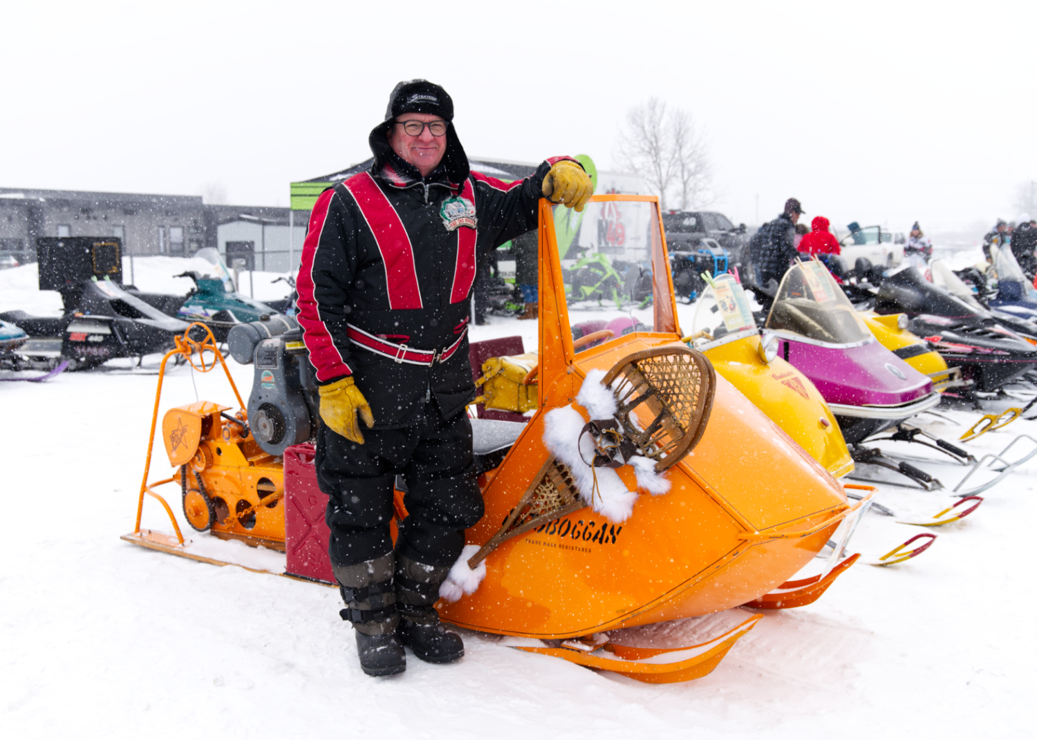 a smiling man stands next to a row of vintage snowmobiles at Big Winter Festiglace on a snowy day.