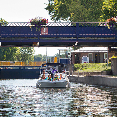 a boat full of happy passengers passes under a bridge surrounded by green trees peaceful buildings on a sunny day. 