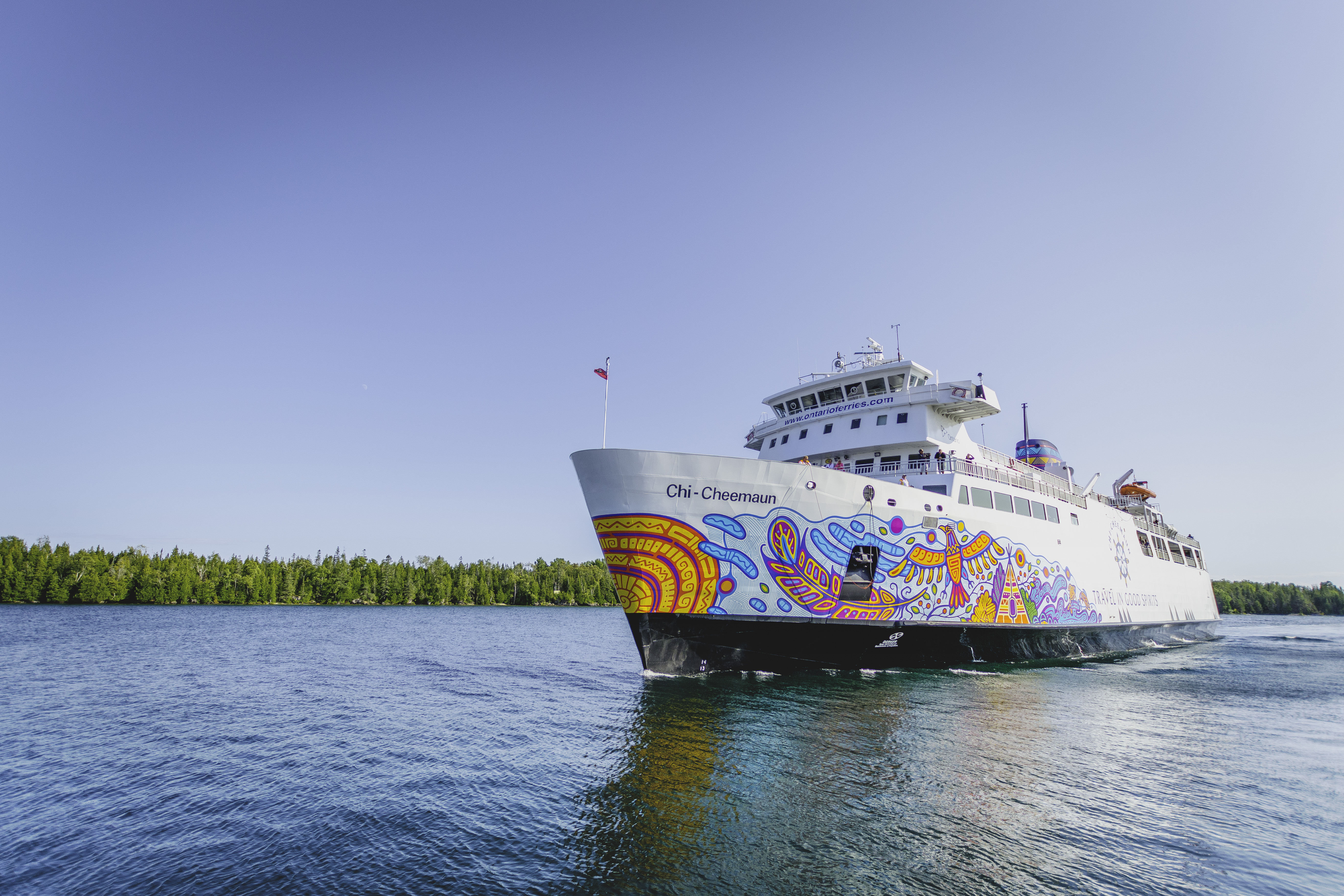 MS Chi Cheemaun; a large white passenger ferry painted with a colourful bird motif sails in the turquoise water of the Georgian Bay under a clear blue sky.