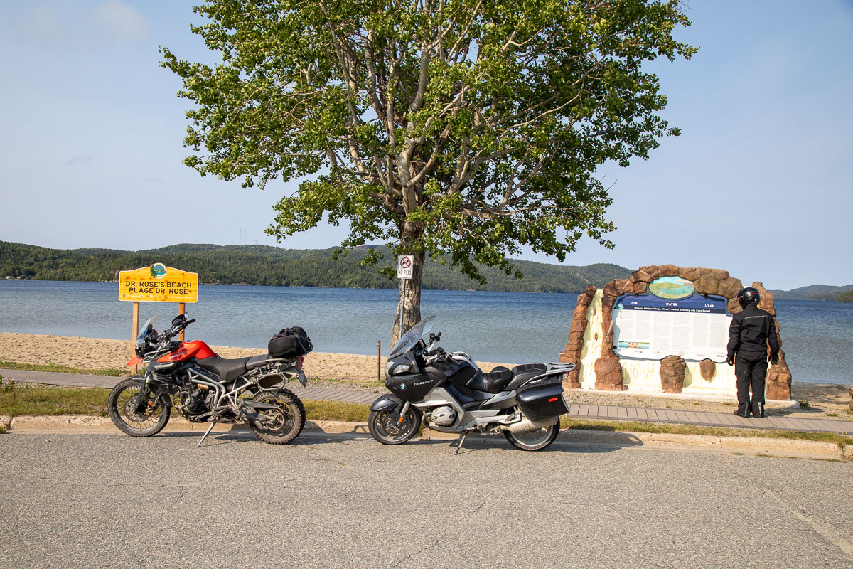 two motorcycles parked next to a lush tree in front of a sandy beach at the edge of a blue lake on a late summer afternoon.