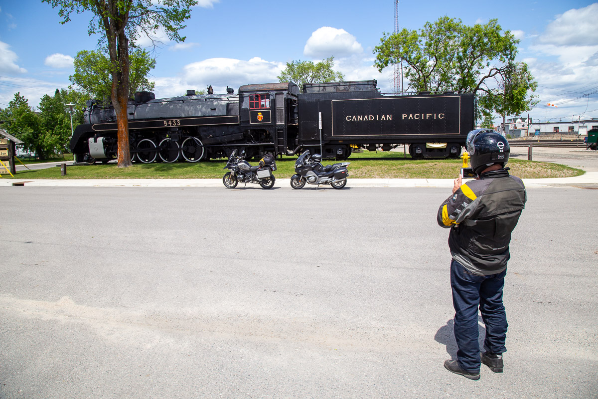 a motorcyclist snaps a photo of a large antique black steam engine on a sunny summer day.