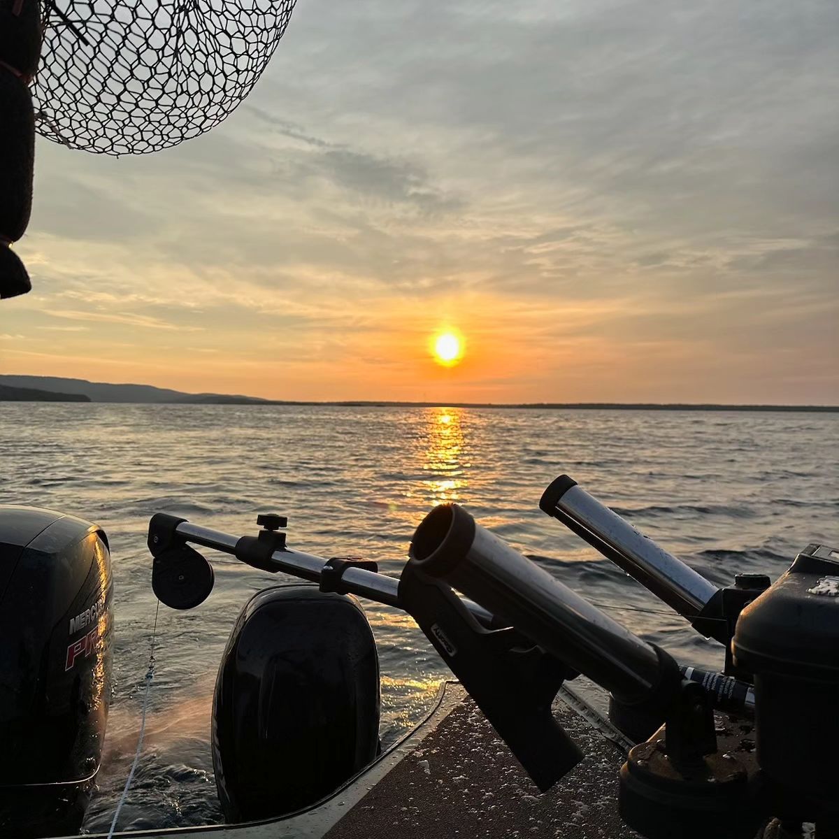 fishing nets and rods against the rail of a motorboat, with shining water illuminated by an orange sunset in the background.