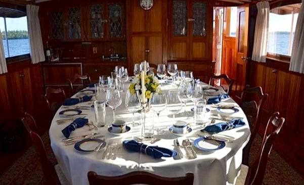 A lavishly laid dining table with china, white linen and stemware in the mohagany cabin of the Grace Anne II ship. Blue lake can be seen through its windows.