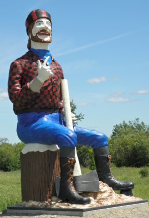 a large wooden statue of a smiling lumberjack sitting on a tree stump with his axe, surrounded by green grass under a blue sky.