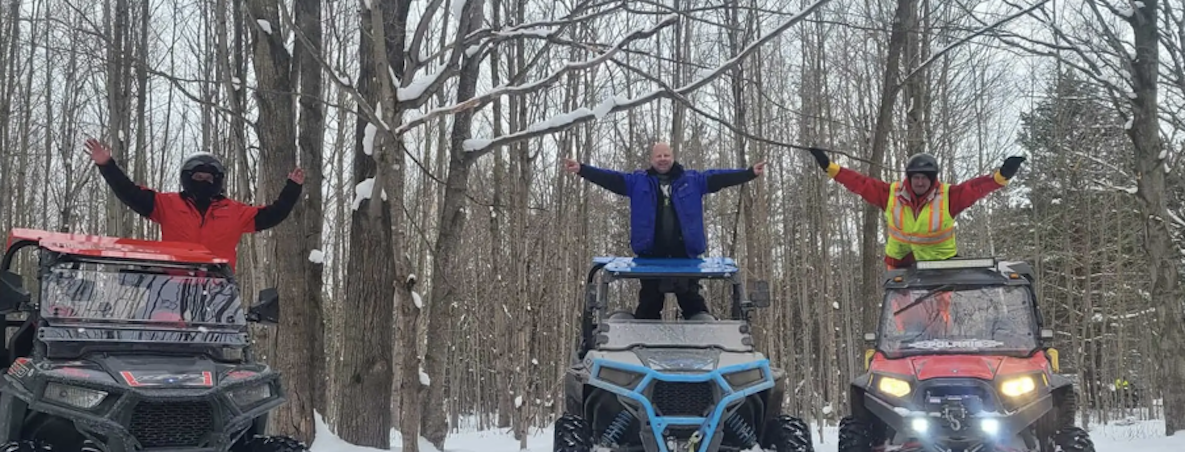 people standing on their ATVs cheering with their arms in the air on a snowy forest trail.