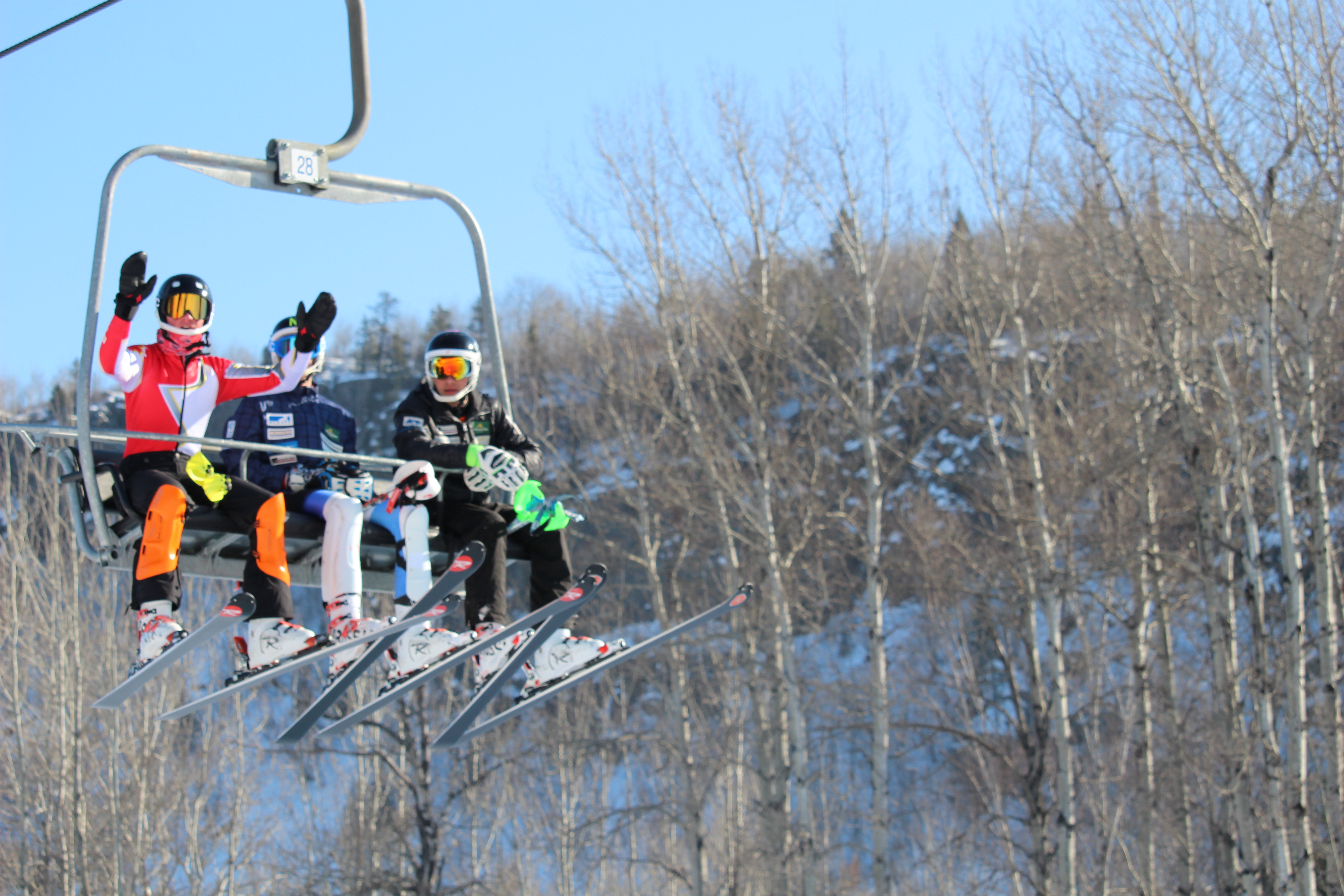 3 people in ski gear wave from a chair lift on a ski hill.