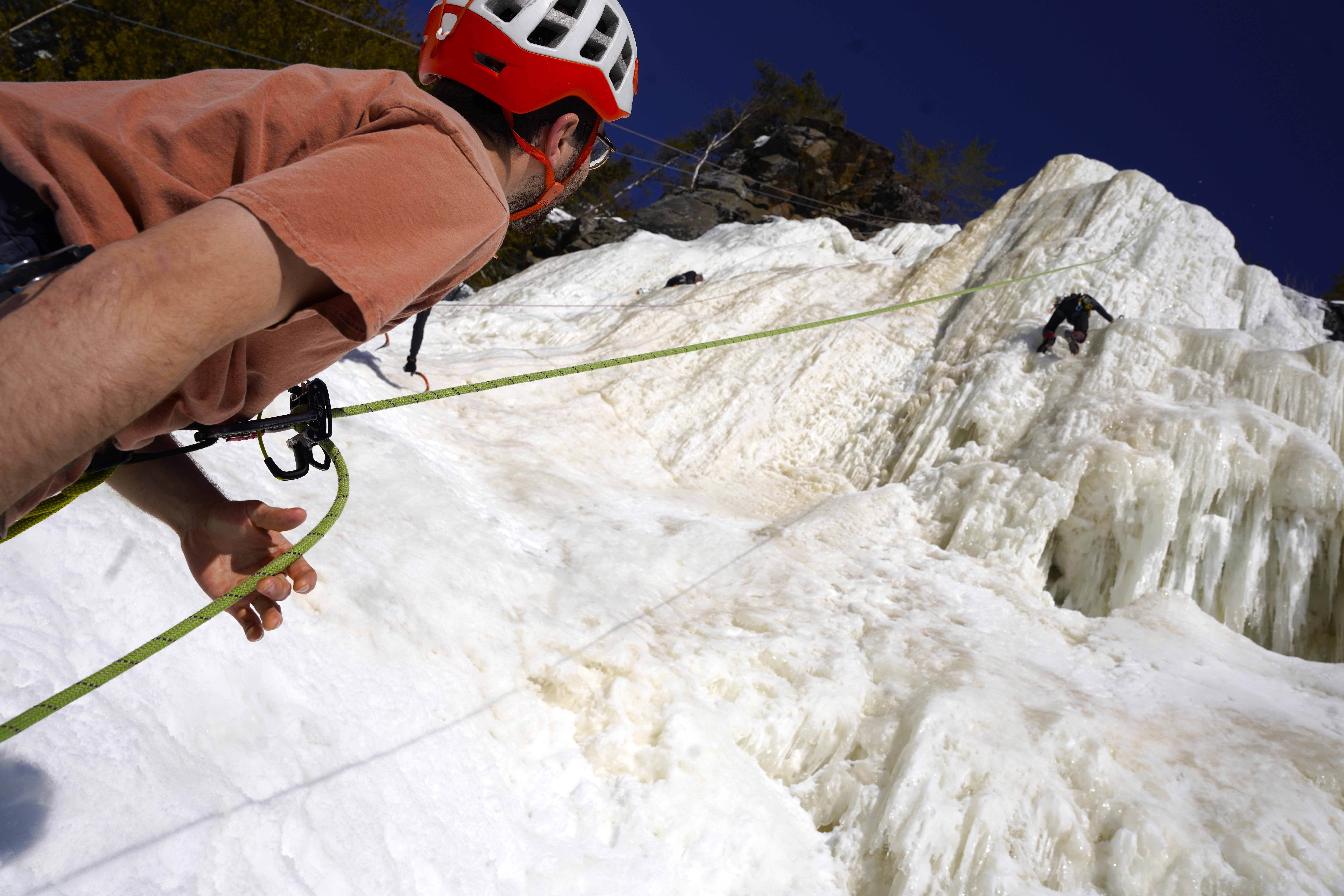 One person at the bottom of an ice wall holds the rope for their partner who can be seen scaling high above on the frozen waterfall.