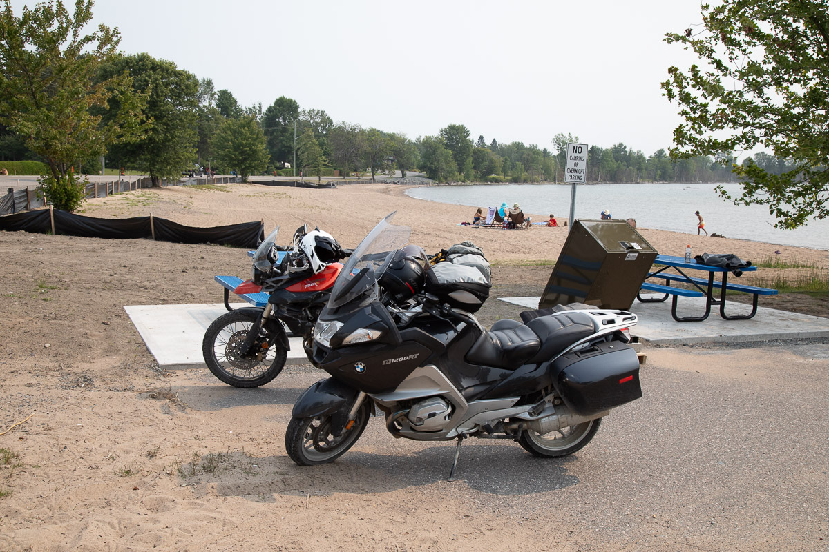 Two motorcycles parked on a sandy beach along a lake's edge on a summer day. 