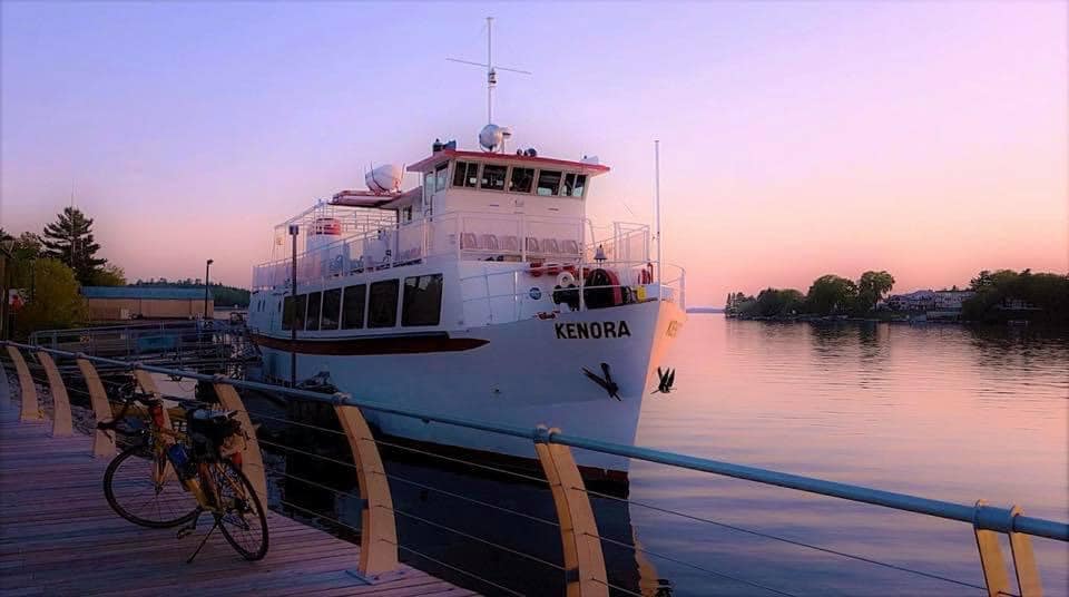 The MS Kenora; a large white cruise ship sits in port on glassy water relecting the pink sunset all around.