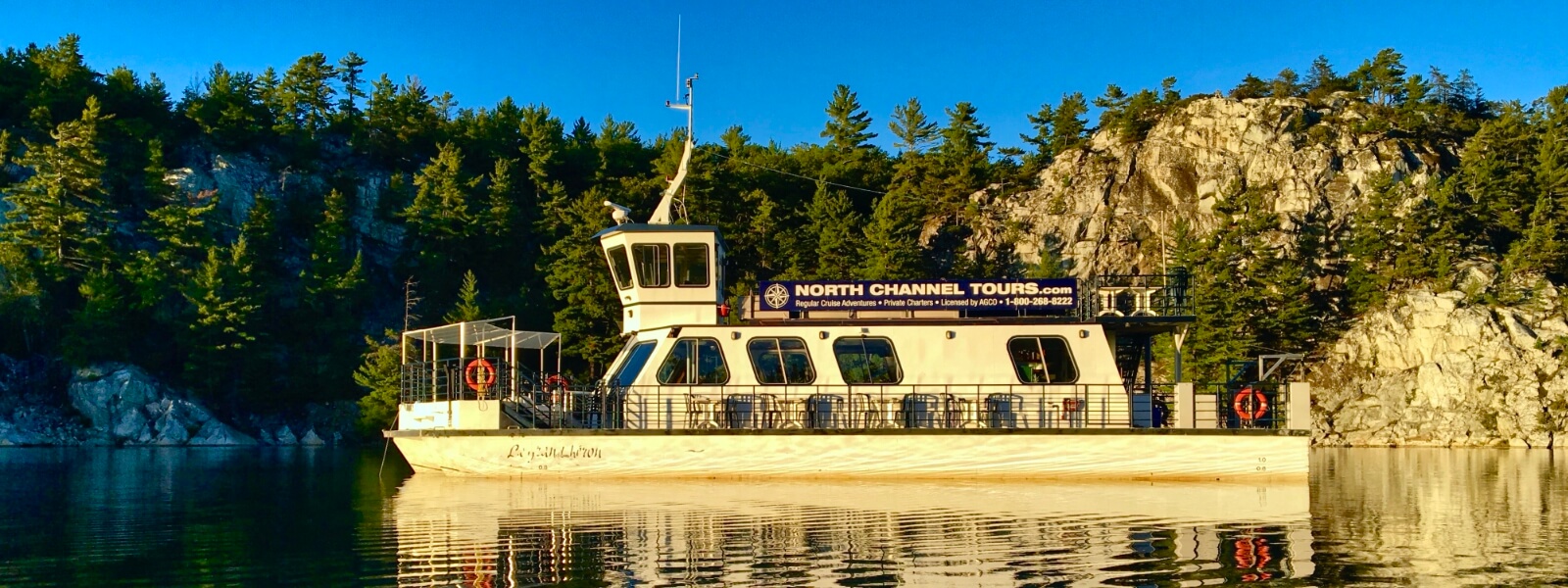 A white cruise boat floats on a glassy lake in the lake afternoon, next to tall rocky banks covered in green trees under a brilliant blue sky.