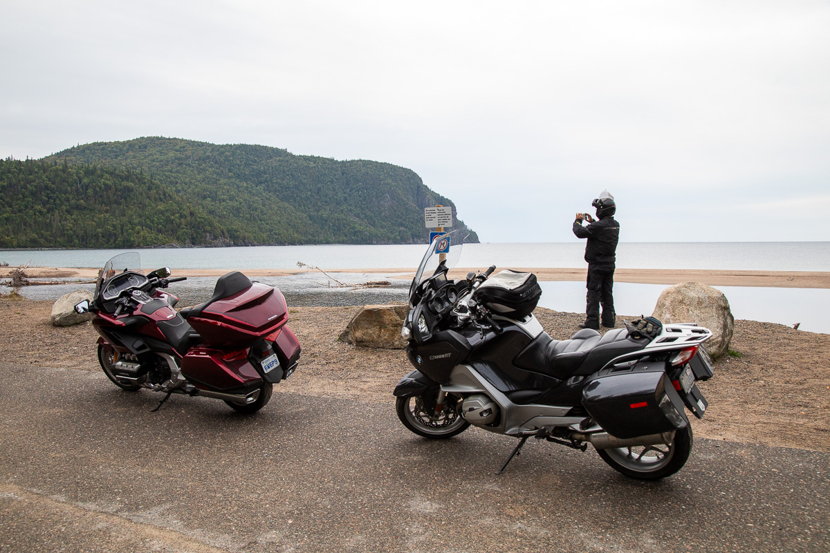 Two motocycles parked at the side of a road while a man in the background takes a photo of the treed, sloping rock formations surrounded by lake water at Old Woman Bay.