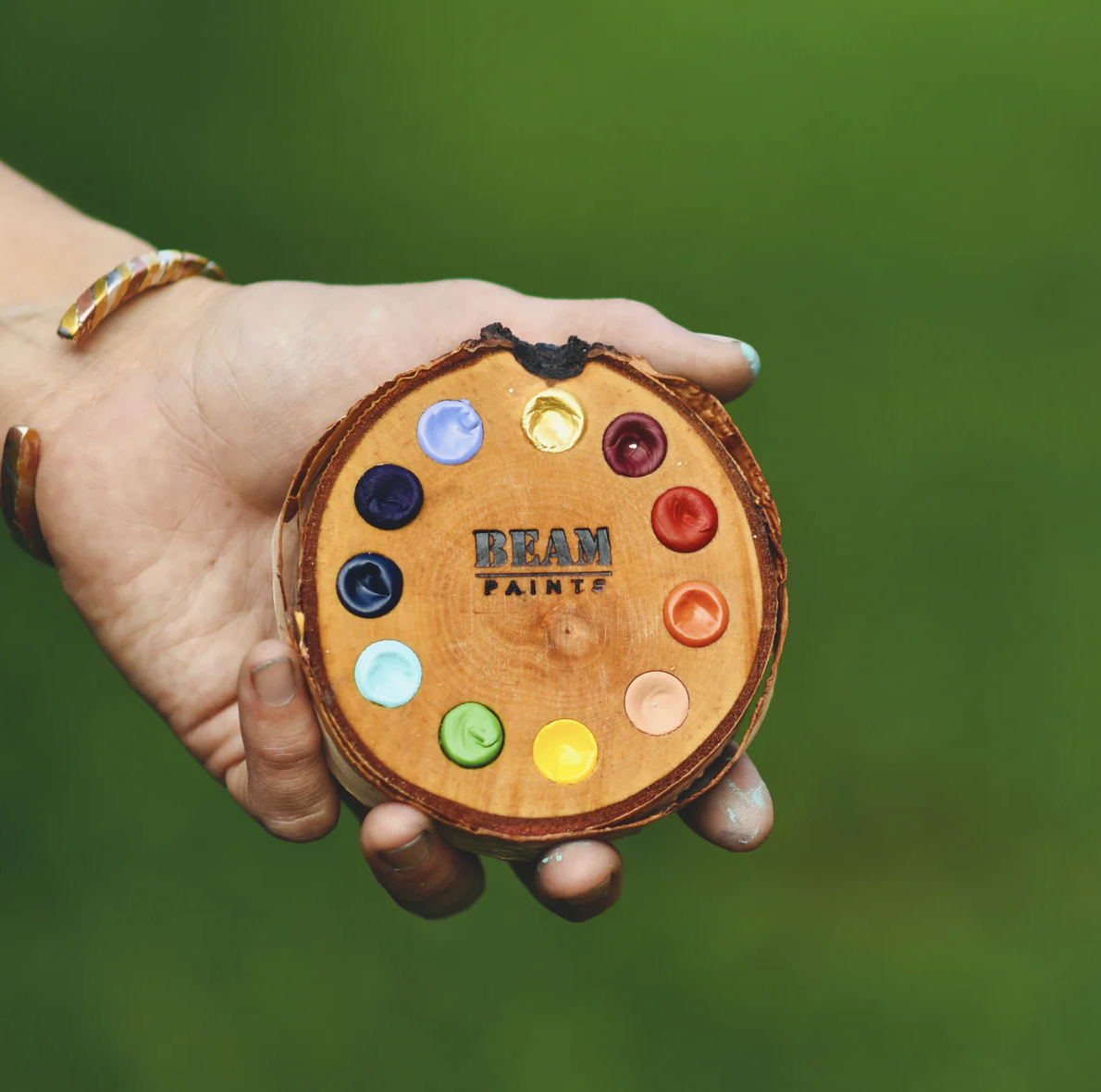 A hand holding a wooden palette filled with many Beam paint colours against a green background.