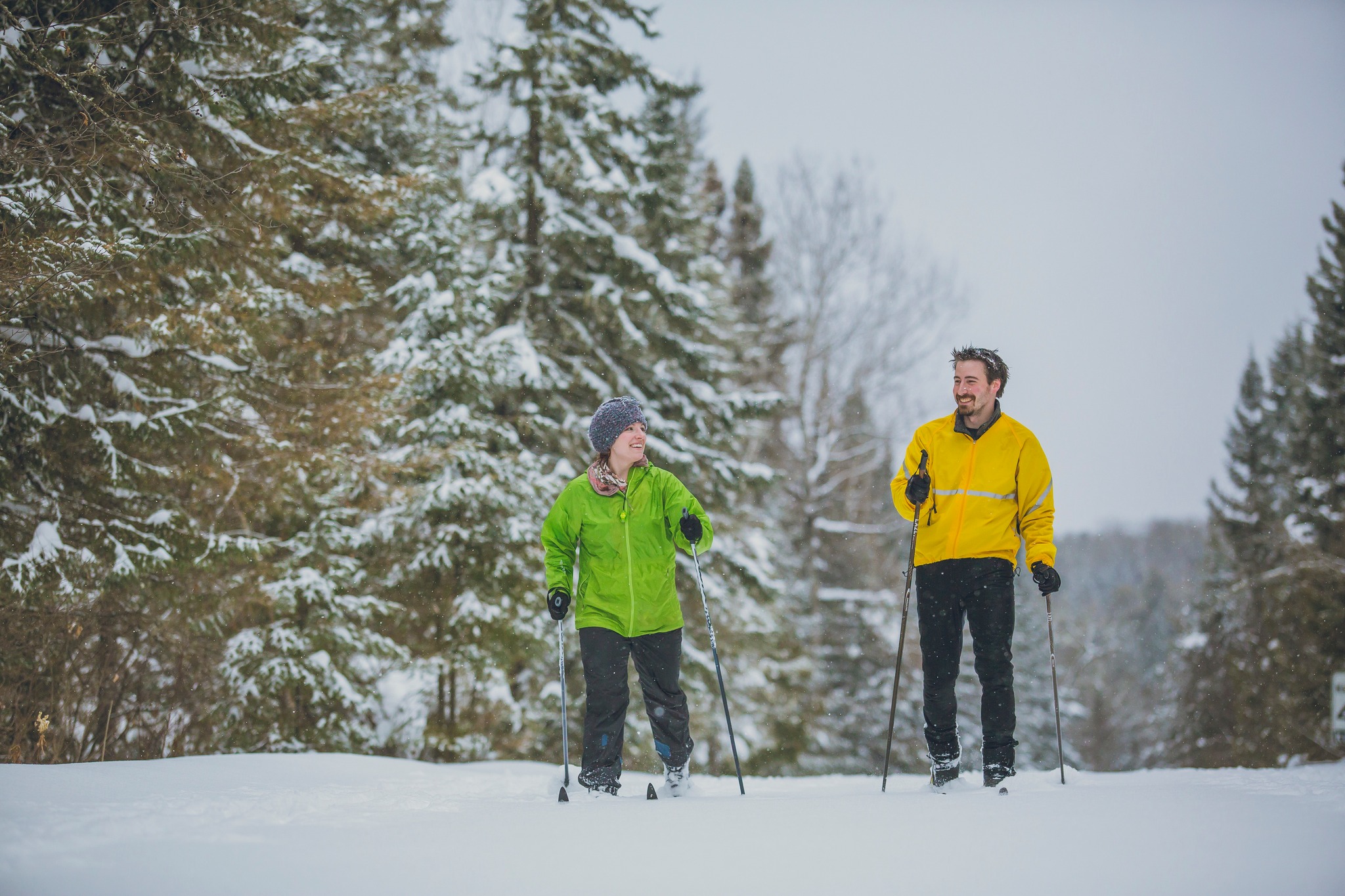 two people smiling and talking cross-country ski through snow-covered forest.