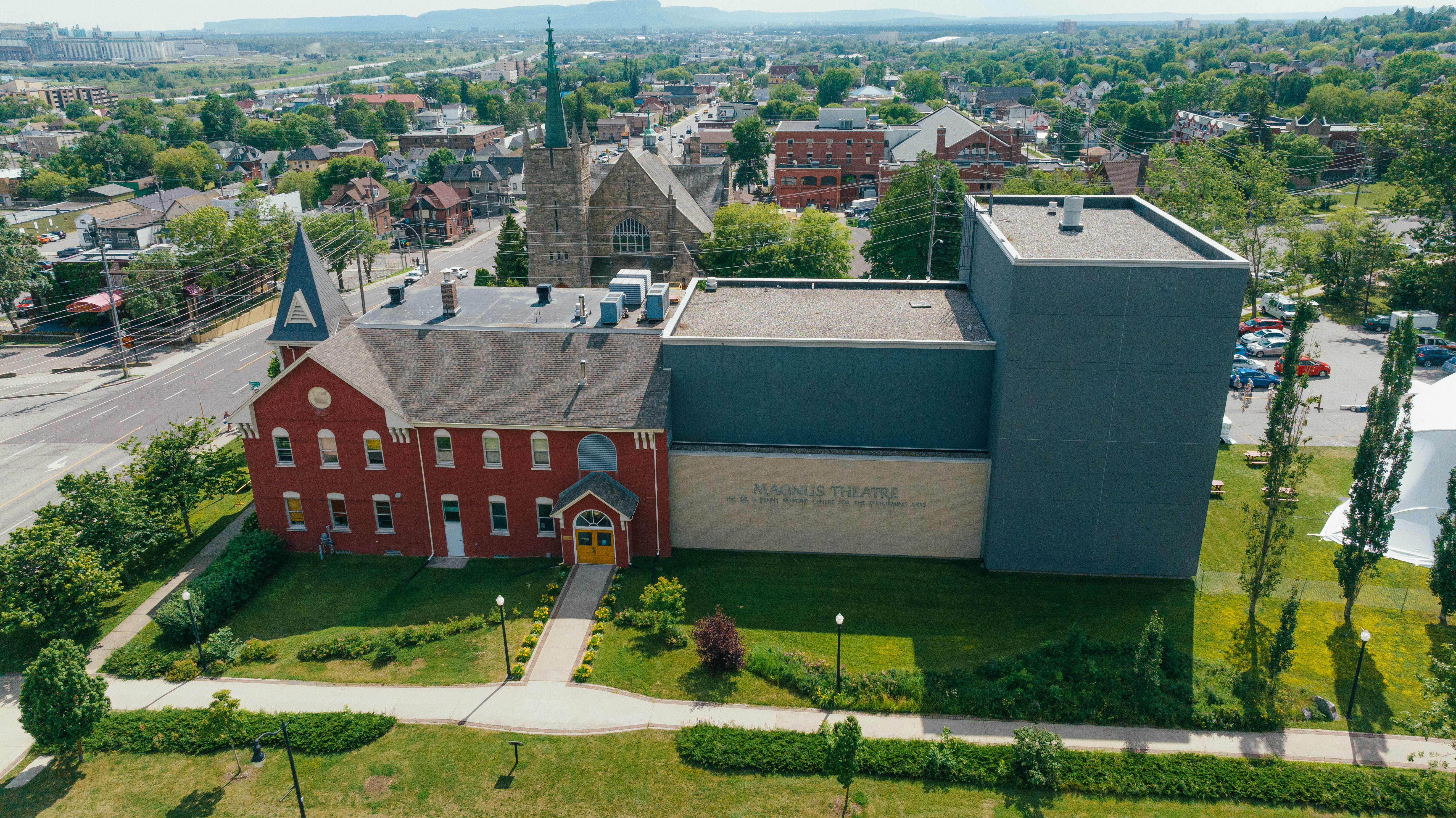 The Magnus Theatre in Thunder Bay; a large, ornate red brick antique building next to a modern grey and white theatre building, surrounded in greenery and the rest of the city of Thunder Bay.