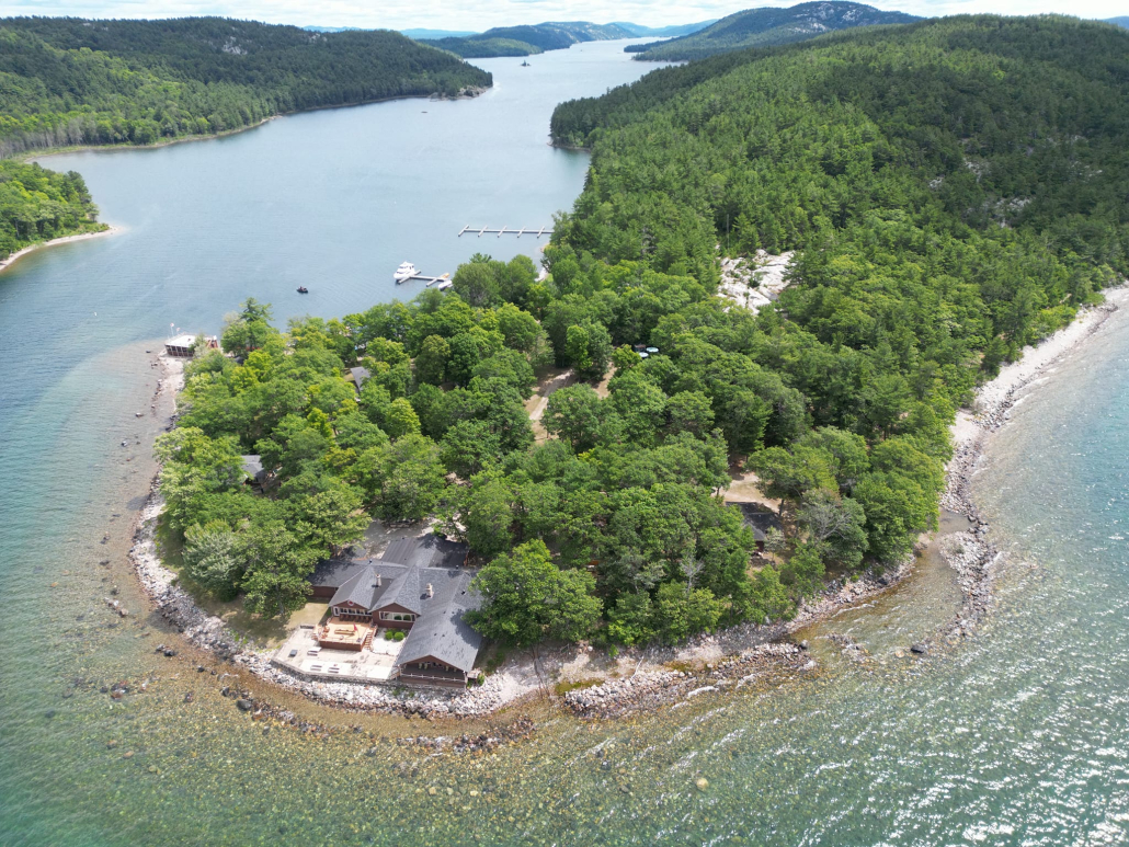 a lush forested island in the middle of clear turquoise water on a sunny summer day. There are lodges and piers nestled among the trees on the water's edge.