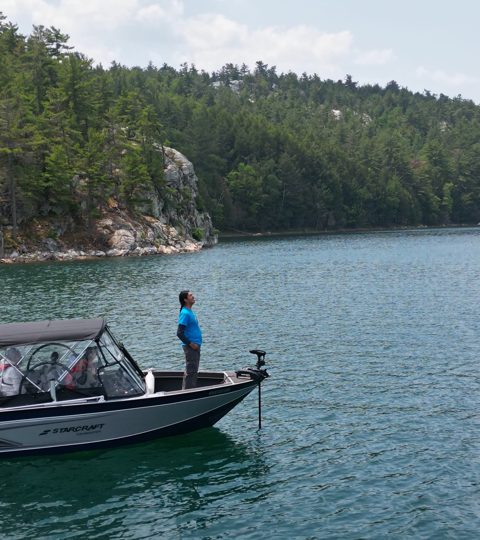 a man standing in the bow of a boat looks up happily at the forested rocky cliffs that surround the lake he's sailing on.