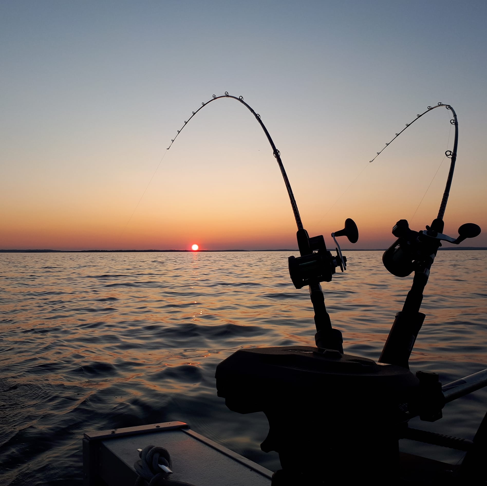 Two fishing rods mounted to the rail of a boat bending under the weight on the line, silhouetted against an orange sunset.