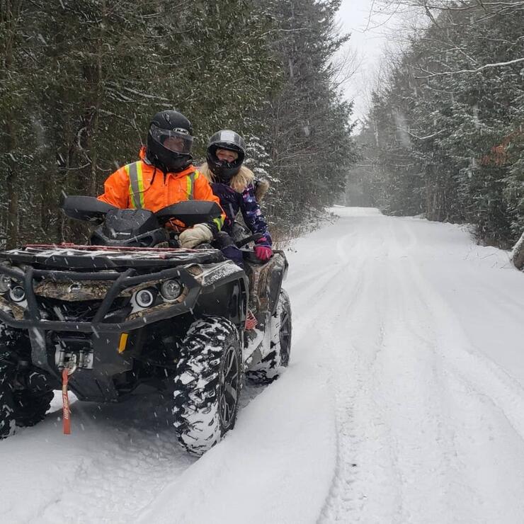 two people ride an ATV on a snowy forest trail in the winter.