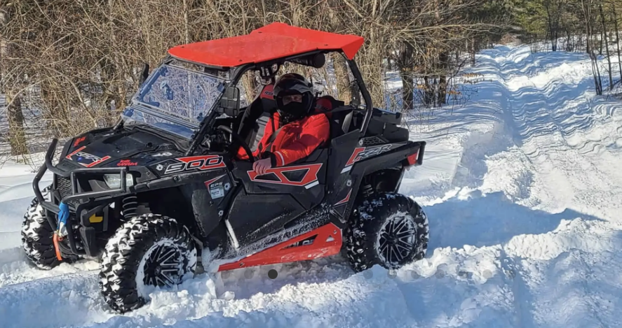 A rider smiles as he rides an ATV down a snowy forest trail on a sunny day.