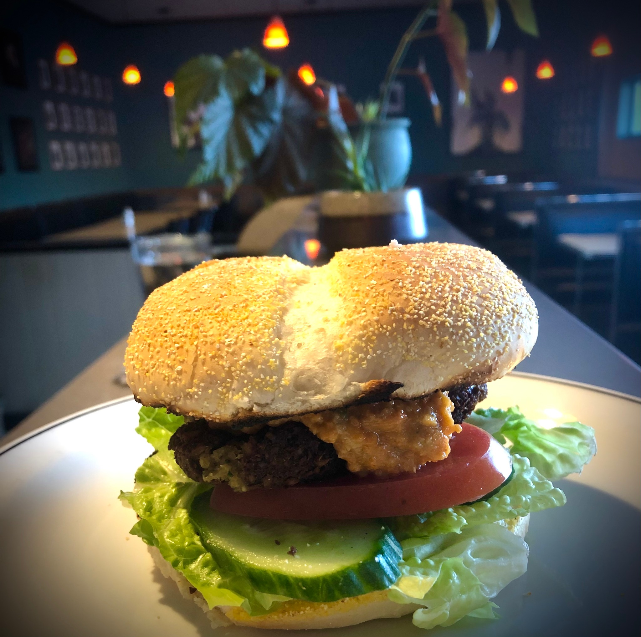 A falafel burger stacked with fresh lettuce, cucumber and tomato under a spotlight. The dining room of Cronos Cafe is in the background. 